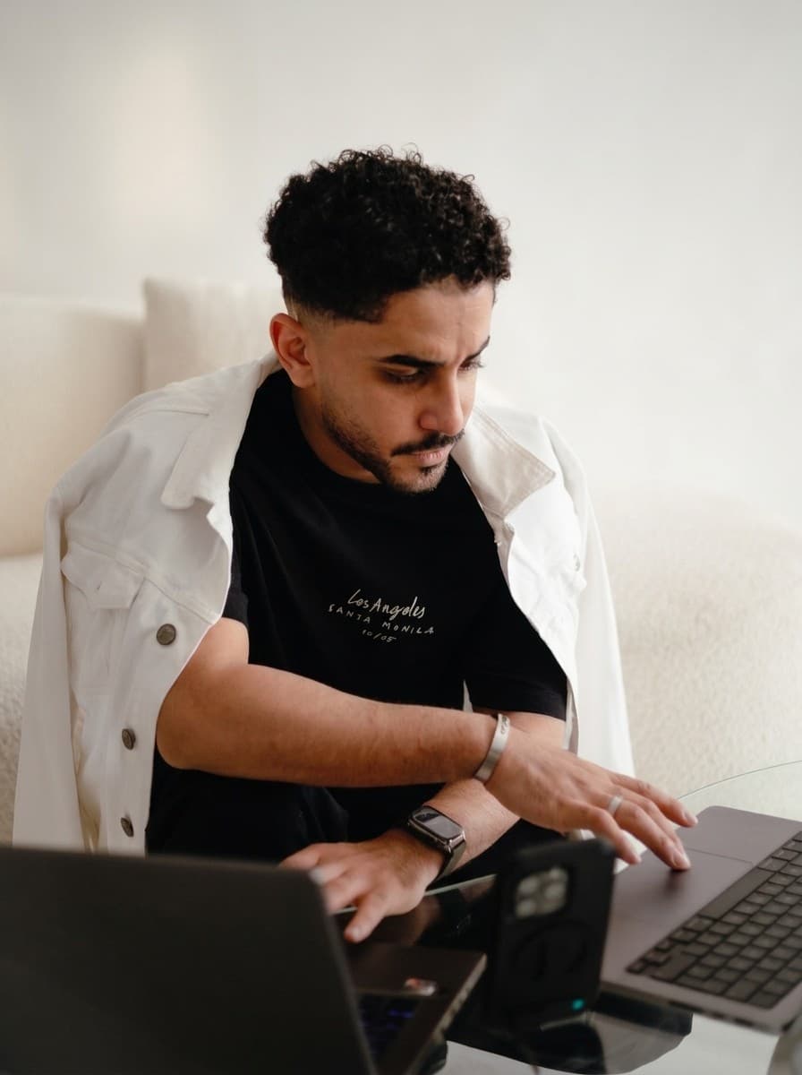 Abdulhadi Alturafi at work on two laptops, in a black t-shirt with a white jacket over his shoulders